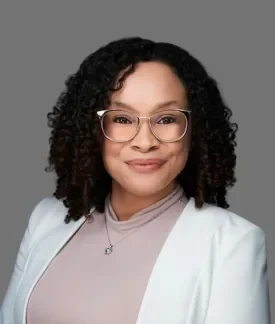 Jordan Gonzalez wearing glasses, a white blazer, and a pink blouse in front of a gray background.