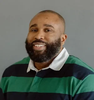 Riche Zamor wearing a blue and green striped rugby shirt with white collar in front of a gray background.