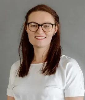 A smiling woman with long dark hair and dark glasses, wearing a white top.