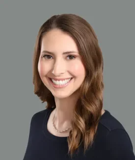 Headshot of white woman with brown hair, smiling, wearing a black shirt in front of a grey background.