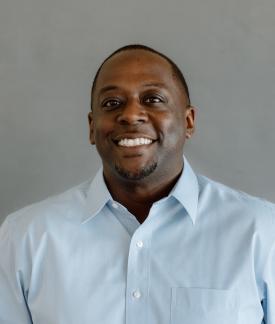 Albert Hughes wearing a blue button down oxford shirt in front of a gray backdrop.
