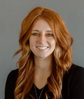 Photo of Alycia Jones, white women with long red hair wearing a black v-neck top in front of a gray background.