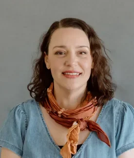 Ana Barcelona wearing a multi-colored scarf and blue shirt in front of a gray background.