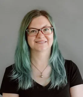 Aubrey Sambor with green hair wearing a black t-shirt in front of a gray background.