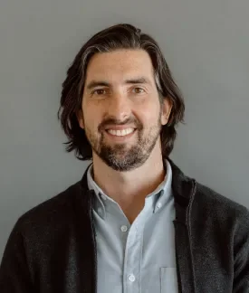 Brian Skowron wearing blue button down shirt with dark gray sweater in front of a gray background.