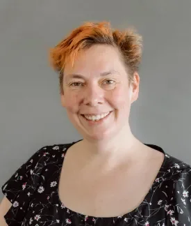 Cathy Theys with orange hair wearing a black blouse with white flowers in front of a gray background.