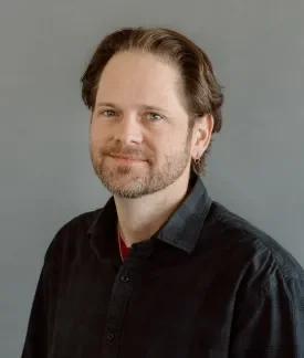 Chris Albrecht wearing a dark button down shirt in front of a gray background.