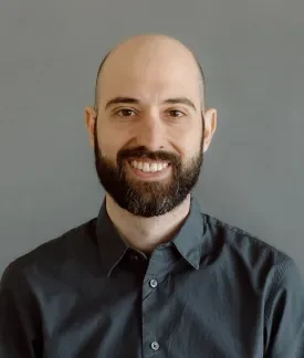 Chris DeLuca wearing a dark button down shirt in front of a gray background.