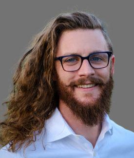 Hawkeye Tenderwolf wearing a blue button down shirt and glasses in front of a gray background.