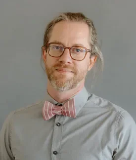 James Sansbury wearing a gray button down shirt with a pink and white striped bowtie in front of a gray background.