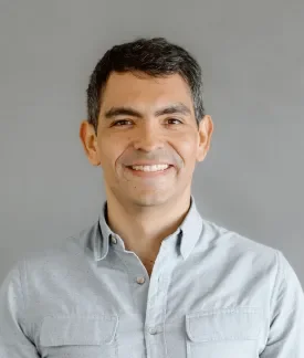 Juampy NR wearing a blue button down shirt standing in front of a gray background.