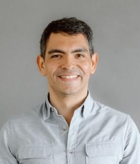 Juampy NR wearing a blue button down shirt standing in front of a gray background.