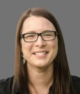 Kris Konrady wearing glasses and wearing a black button down shirt in front of a gray background.