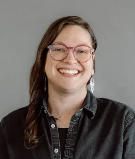 Marissa Epstein wearing glasses and a black button down shirt in front of a gray background.
