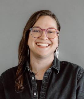 Marissa Epstein wearing glasses and a black button down shirt in front of a gray background.