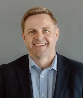 Matt Kleve wearing a blue button down shirt with a navy sport coat in front of a gray background.