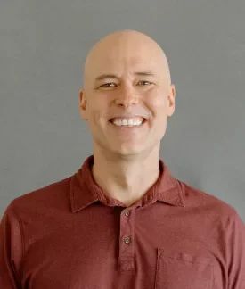 Matthew Tift wearing a red button down shirt in front of a gray background.
