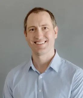 Nate Lampton wearing a blue biutton down shirt in front of a gray background.