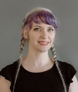 Pauline Judge wearing a short-sleeved black shirt and braided pigtails with purple hair in front of a gray background.