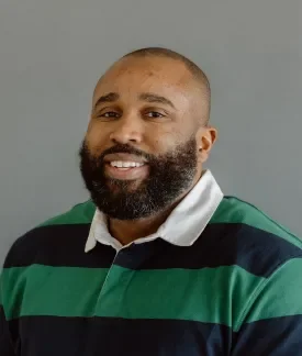 Riche Zamor wearing a blue and green striped rugby shirt with white collar in front of a gray background.