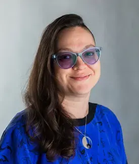 Marissa Epstein is a woman in her mid-30s with long brown hair shaved on one side, wearing a purple and black blouse, purple cat-eye glasses, a silver necklace, and a smirk.