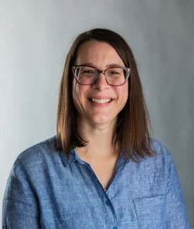 A smiling woman with straight brown hair and glasses, wearing a light blue button-up shirt, standing in front of a neutral gray background.