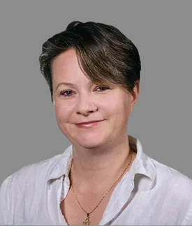 Professional headshot of a woman with short, side-swept brown hair and fair skin, smiling gently at the camera. She is wearing a white button-down shirt and a delicate gold necklace, posed against a plain gray background.