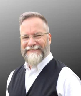 Portrait of a smiling man with short gray hair, a full gray beard, and round glasses, wearing a white button-down shirt and dark vest against a neutral gray background.
