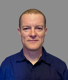A person with a closely cropped haircut and small hoop earrings stands facing the camera against a plain light-gray wall, wearing a dark navy button-up shirt and a slight smile.