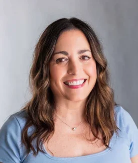 Woman with long, wavy brown hair, brown eyes, wearing a light blue scoop-neck t-shirt in front of a gray background.