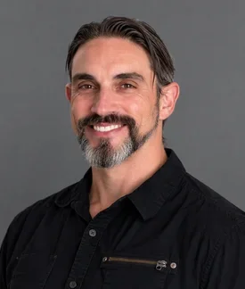 Smiling man with dark, graying hair and a salt-and-pepper beard, wearing a black button-up shirt against a medium-gray background.