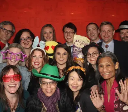 A group of thirteen Lullabot employees pose in a photo booth wearing an assortment of goofy hats and glasses.