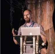 Jared stands on a stage in front of a podium with his laptop and gestures while he talks.