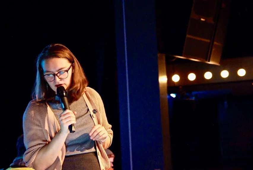 Sally stands on a stage holding a microphone to her mouth as she speaks.