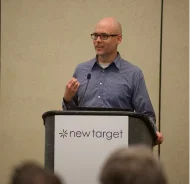 Matthew stands on a stage in front of a podium with a sign that reads, new target.