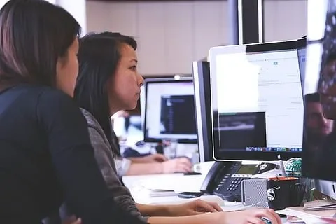 Two people sitting at a desk, both looking at the same computer monitor.