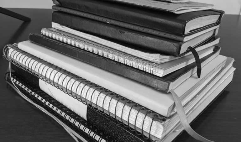 a dozen old used sketchbooks piled on a desk, in black and white 