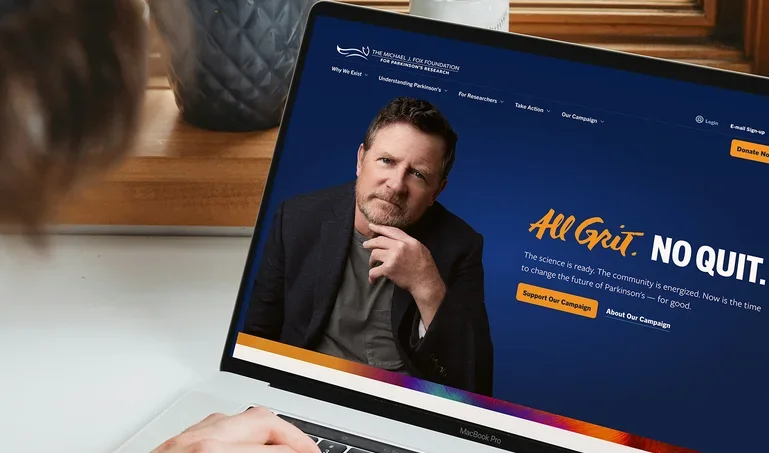 A person using a laptop displaying The Michael J. Fox Foundation for Parkinson’s Research website, featuring the “All Grit. No Quit.” campaign with a prominent image of Michael J. Fox and links to support the campaign.