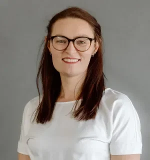 A smiling woman with long dark hair and dark glasses, wearing a white top.