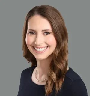 Headshot of white woman with brown hair, smiling, wearing a black shirt in front of a grey background.