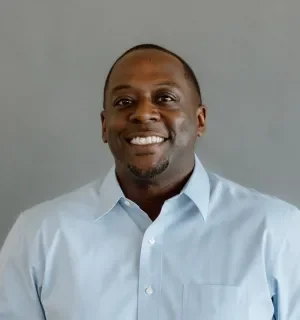 Albert Hughes wearing a blue button down oxford shirt in front of a gray backdrop.
