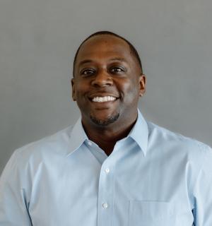 Albert Hughes wearing a blue button down oxford shirt in front of a gray backdrop.