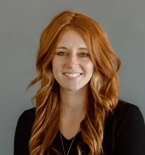 Photo of Alycia Jones, white women with long red hair wearing a black v-neck top in front of a gray background.