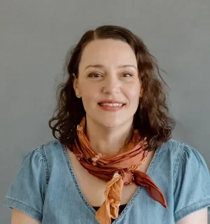 Ana Barcelona wearing a multi-colored scarf and blue shirt in front of a gray background.