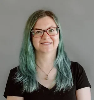 Aubrey Sambor with green hair wearing a black t-shirt in front of a gray background.