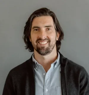 Brian Skowron wearing blue button down shirt with dark gray sweater in front of a gray background.