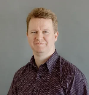 Bryan Heisler wearing a dark button down shirt with tiny polka dots in front of a gray background.