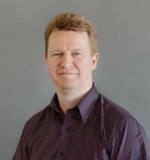 Bryan Heisler wearing a dark button down shirt with tiny polka dots in front of a gray background.