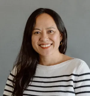 Cat Rude wearing a long-sleeved white sweater with black stripes in front of a gray background.