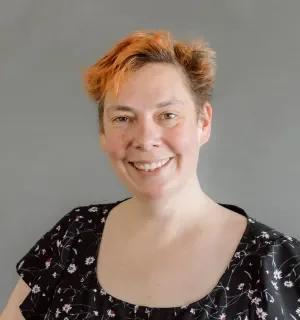 Cathy Theys with orange hair wearing a black blouse with white flowers in front of a gray background.